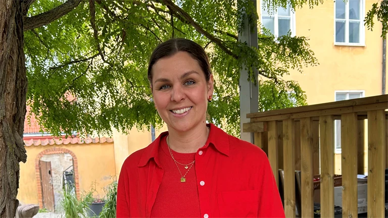 Lina Ejlertsson står under ett lövträd och ser in i kameran. Hon har på sig en röd tröja, röd skjorta och vita byxor.