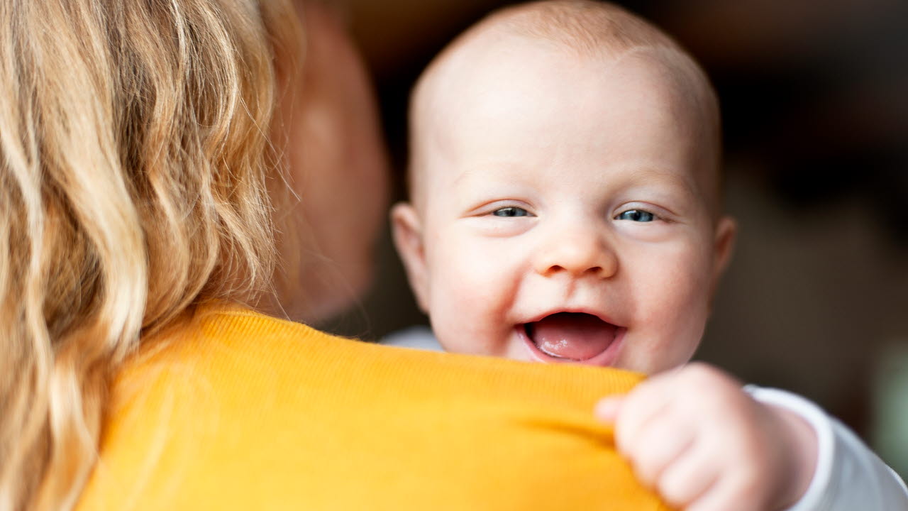 Kvinna håller ett litet barn i famnen. Barnet tittar mot kameran och ler stort.