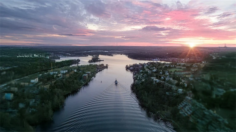 En flod i en fjärd omgiven av skog sedd uppifrån, i solnedgången, med rosa och lila moln på ljus himmel. Redigerad med AI: