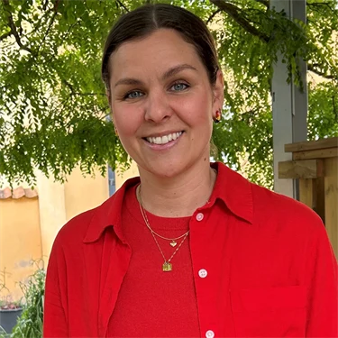 Lina Ejlertsson står under ett lövträd och ser in i kameran. Hon har på sig en röd tröja, röd skjorta och vita byxor.