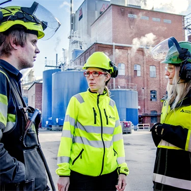 Tre personer i varselkläder och skyddshjälmar står utomhus framför Aspa bruk. Det ryker ur skorstenarna.