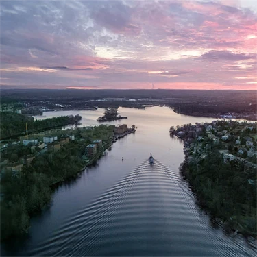 En flod i en fjärd omgiven av skog sedd uppifrån, i solnedgången, med rosa och lila moln på ljus himmel. Redigerad med AI: