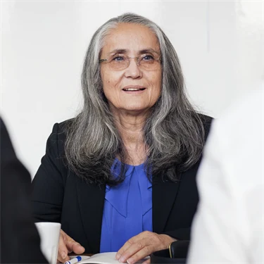 Äldre kvinna i långt, grått hår och glasögon sitter i möte med två andra personer.