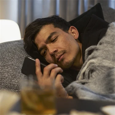 Sjuk man ligger under en filt i soffan och kollar mobilen.