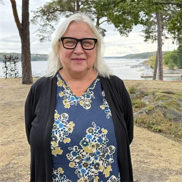 Forskare Maria Åkesson har ljust hår, glasögon med mörka bågar, en mellanblå blus med blommiga mönster och en mörkblå kofta, i bakgrunden syns utsikt över vatten och grönområden.