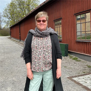 Gunnel Hensing forskare vid Göteborgs universitet står framför ett rödmålat trähus, hon har glasögon, ljusblå jeans, blommig tröja, scarf och en grå lång kofta på sig
