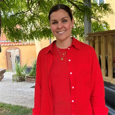 Lina Ejlertsson står under ett lövträd och ser in i kameran. Hon har på sig en röd tröja, röd skjorta och vita byxor.