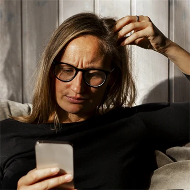 Kvinna sitter i soffan och tittar på sin mobiltelefon med bekymrad min.