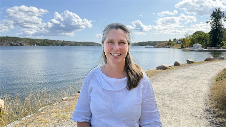 Forskare Eva Lindell har en vit blus och ler mot kameran, i bakgrunden syns utsikt över vatten och grönområden.