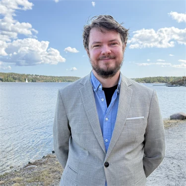 Forskare Andreas Bergqvist har skägg, ljusblå skjorta och ljusgrå kavaj, i bakgrunden syns utsikt över vatten och grönområden.