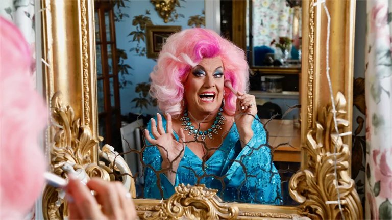 Babsan iklädd rosa peruk skrattar och tittar in i en spegel med guldram.