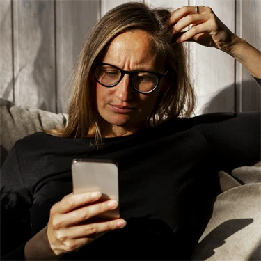 Kvinna sitter i soffan och tittar på sin mobiltelefon med bekymrad min.