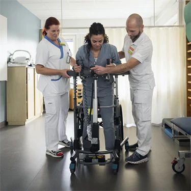 En person som rehabilteras. Hen står i mitten och har två sjukvårdare bredvid sig.