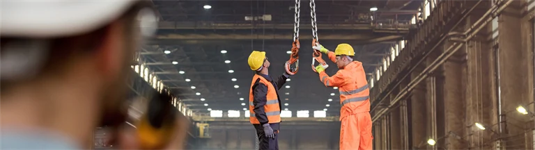 Två män i arbetskläder och hjälm står i fabrik och fixar med hängande kedja från taket