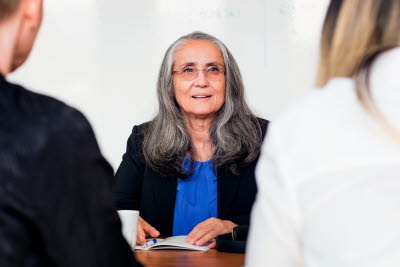Äldre kvinna i långt, grått hår och glasögon sitter i möte med två andra personer.