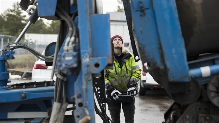 Kvinna i arbetskläder manövrerar stor maskin eller fordon med joystick eller liknande,