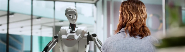 En robot syns framifrån vid ett kontorsbord gestikulerar mot en kvinna som sitter mittemot, med ryggen mot.