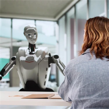 En robot syns framifrån vid ett kontorsbord gestikulerar mot en kvinna som sitter mittemot, med ryggen mot.