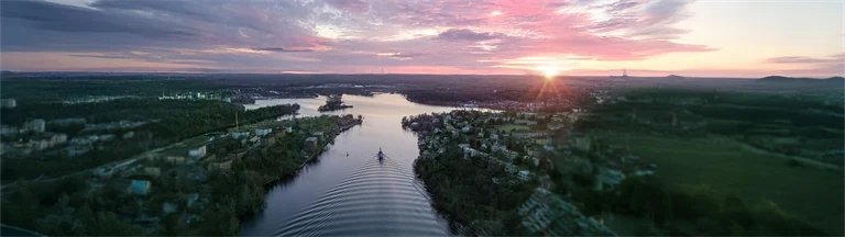 En flod i en fjärd omgiven av skog sedd uppifrån, i solnedgången, med rosa och lila moln på ljus himmel. Redigerad med AI: