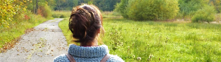 Kvinna med uppsatt hår och ryggsäck går på en stig mot en höstfärgad skog.