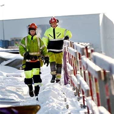 Personer i varselkläder och hjälm jobbar utomhus i ett snöigt vinterlandskap.