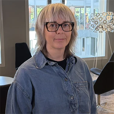 Forskare Åsa Tjulin har ljust hår, glasögon med mörka bågar, blå skjorta och mörka byxor, hon står i en loungemiljö