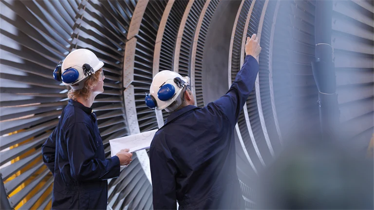 Två ingenjörer i hjälmar och blå arbetsställ inspekterar en en turbin.