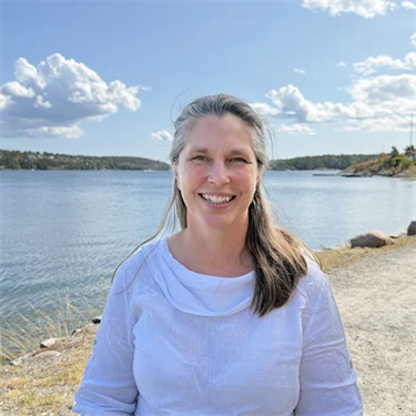 Forskare Eva Lindell har en vit blus och ler mot kameran, i bakgrunden syns utsikt över vatten och grönområden.
