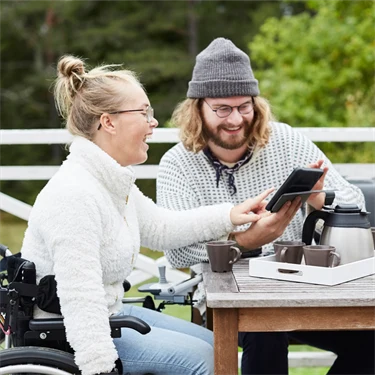 Manlig vårdgivare och funktionshindrad kvinna sitter vid bord på altan och använder surfplatta tillsammans