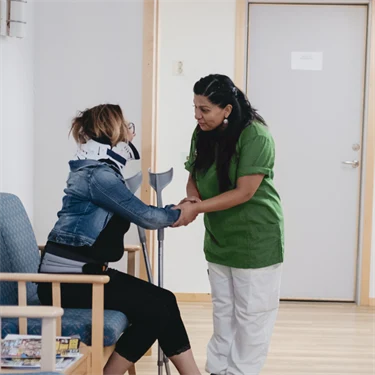 Skadad kvinna med nackkrage och kryckor sitter i väntrum och hälsar på sjuksköterska