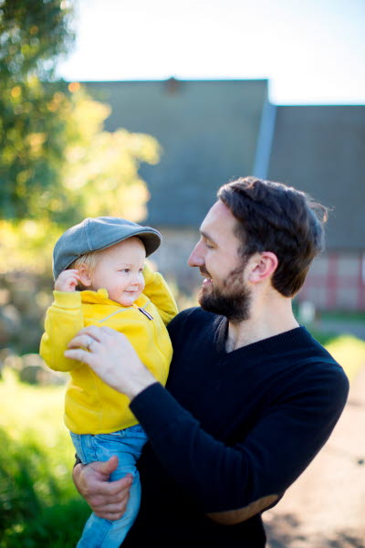 Skäggig man håller ett småbarn med gul jacka i famnen
