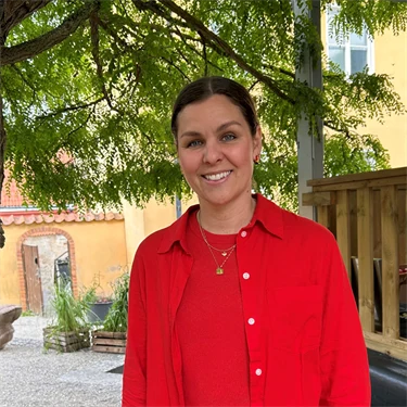 Lina Ejlertsson står under ett lövträd och ser in i kameran. Hon har på sig en röd tröja, röd skjorta och vita byxor.