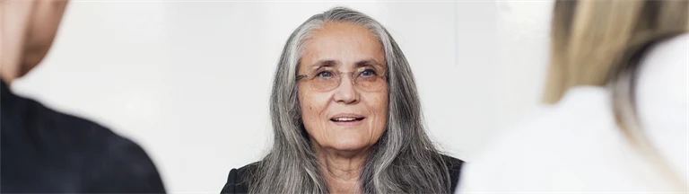 Äldre kvinna i långt, grått hår och glasögon sitter i möte med två andra personer.