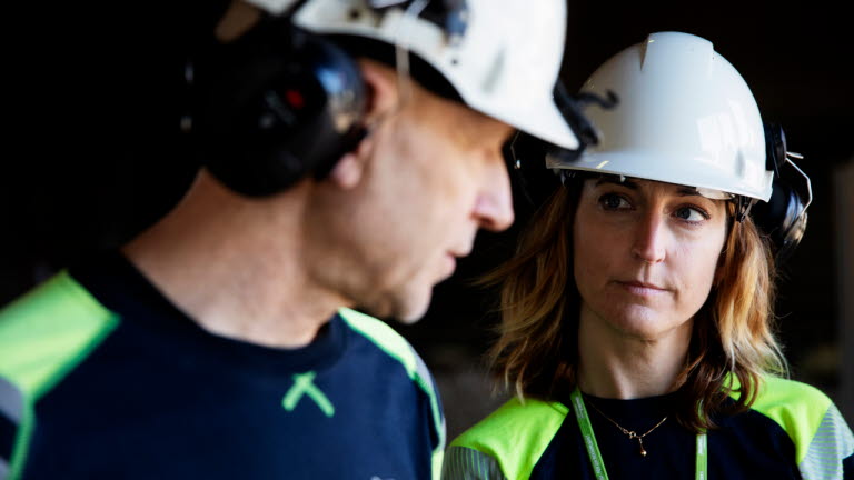 Man och kvinna med skyddshjälmar samtalar i fabrik i Sandviken.
