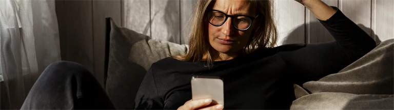 Kvinna sitter i soffan och tittar på sin mobiltelefon med bekymrad min.
