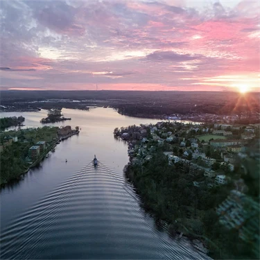 En flod i en fjärd omgiven av skog sedd uppifrån, i solnedgången, med rosa och lila moln på ljus himmel. Redigerad med AI: