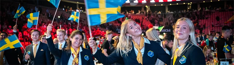 Några av ungdomarna i yrkeslandslaget ler och viftar med svenska flaggor vid middagsbordet under invigningen av Yrkes-EM.