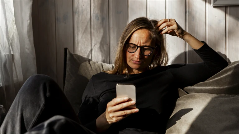 Kvinna sitter i soffan och tittar på sin mobiltelefon med bekymrad min.
