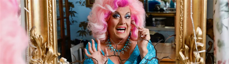 Babsan iklädd rosa peruk skrattar och tittar in i en spegel med guldram.