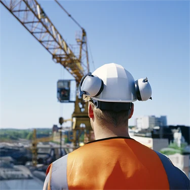 Manlig byggnadsarbetare med hjälm och hörselskydd blickar ut över lyftkran