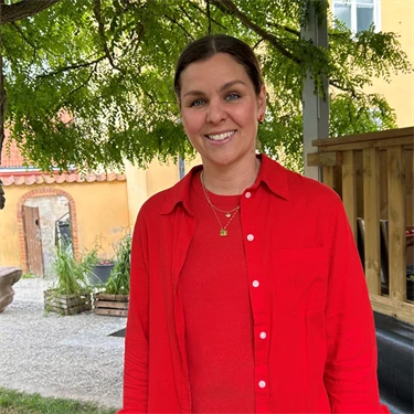 Lina Ejlertsson står under ett lövträd och ser in i kameran. Hon har på sig en röd tröja, röd skjorta och vita byxor.