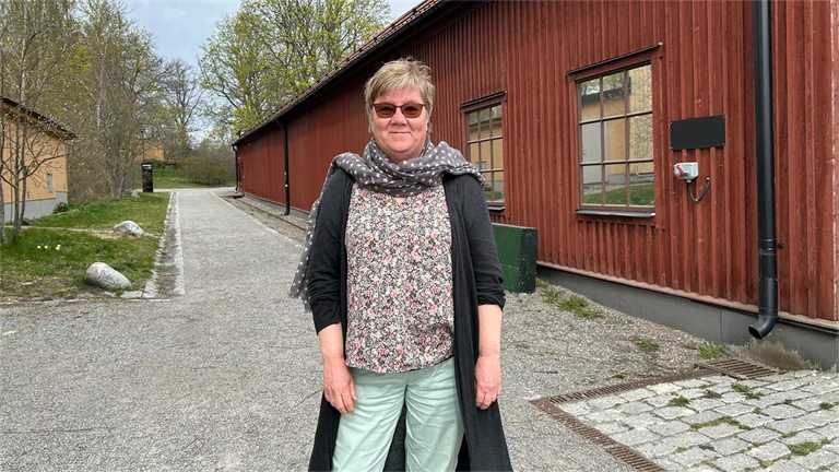Gunnel Hensing forskare vid Göteborgs universitet står framför ett rödmålat trähus, hon har glasögon, ljusblå jeans, blommig tröja, scarf och en grå lång kofta på sig