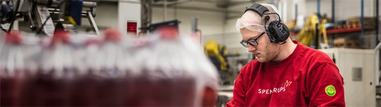 En person arbetar i produktionen i Spendrups fabrik. Personen har en röd tröja, glasögon och hörselkåpor på sig. 