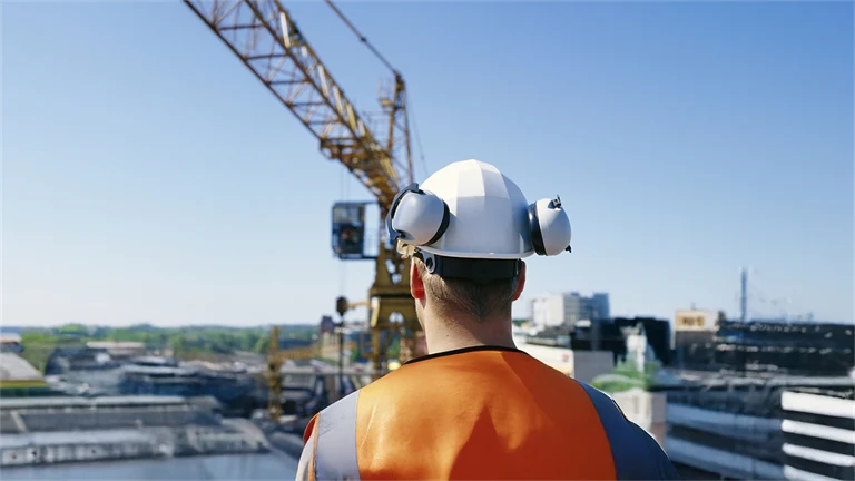 Manlig byggnadsarbetare med hjälm och hörselskydd blickar ut över lyftkran