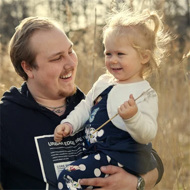 Stålverksarbetaren Emil Heed har sin dotter i famnen. De är utomhus och plockar olika strån.