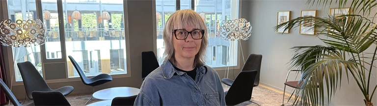 Forskare Åsa Tjulin har ljust hår, glasögon med mörka bågar, blå skjorta och mörka byxor, hon står i en loungemiljö