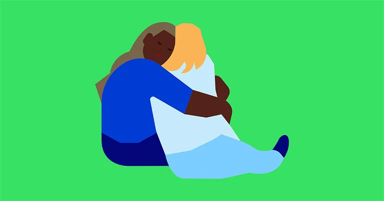 Kvinna tröstar en annan med en kram, de sitter på golvet.
