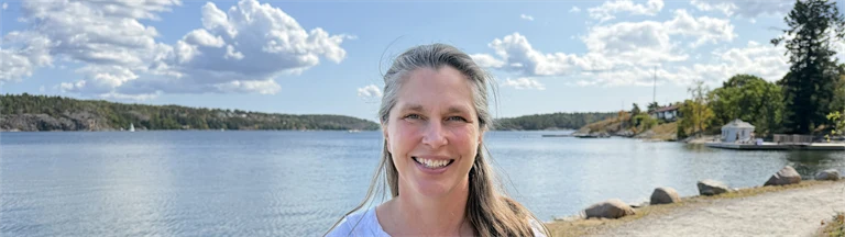 Forskare Eva Lindell har en vit blus och ler mot kameran, i bakgrunden syns utsikt över vatten och grönområden.