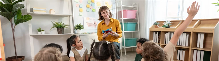 En förskolepedagog står framför en barngrupp. Pedagogen ler och håller upp ett flygplan, ett barn räcker upp handen. 