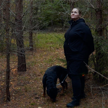 Djursjukskötaren Martina Sandrén rastar sin hund i en skog med tallar och granar. Hunden står bredvid henne och det är grön mossa på marken och solljus skiner in mellan träden.